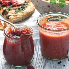 Dwa słoiki domowego sosu do pizzy, jeden otwarty z łyżką pełną aromatycznej masy, obok kawałek pizzy i przyprawy.