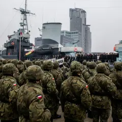 Żołnierze w mundurach polowych stoją w szyku przed okrętem wojennym. W tle nowoczesne budynki i inni wojskowi. To część ćwiczeń wojskowych.