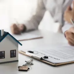 Maquette de maison, clés et contrat sur une table. Des mains signent des documents, symbolisant le **délai achat maison**.
