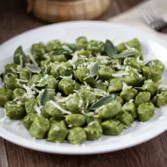 Zielone kopytka ze szpinakiem, posypane parmezanem i listkami szałwii, prezentują się apetycznie na białym talerzu.