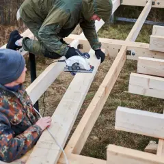 Dwóch mężczyzn buduje konstrukcję z drewna. Jeden z nich używa piły tarczowej do cięcia belek, które są częścią projektu.