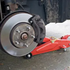 Hamulec samochodowy z tarczą i zaciskiem, gotowy do wymiany. Czerwony podnośnik hydrauliczny, jak jarzmo, czeka na swoją kolej.