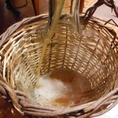 Płyn przelewany do szklanej butli w wiklinowym koszu, tworząc pianę.