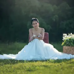 Uśmiechnięta panna młoda w białej sukni siedzi na krześle w ogrodzie. Jej tren rozpościera się na trawie.