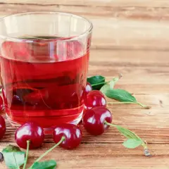 Szklanka z rubinowym kompotem z wiśni na zimę, otoczona świeżymi owocami i listkami na drewnianym tle.