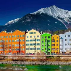 Colorful buildings line the Inn River, a vibrant scene on the journey from St. Moritz to Innsbruck, with snow-capped mountains in the background.