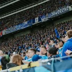 Tłum kibiców na stadionie, w niebieskich barwach, z transparentami "Jaworzno", "Mikołów", "Chorzów". Wszyscy w zgodzie dopingują swoją drużynę.