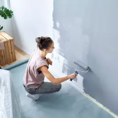 Frau malt Wand grau mit Rolle, um sie streifenfrei zu streichen. Möbel sind abgedeckt.