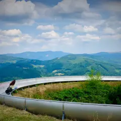 Dwie osoby zjeżdżają na sankach po torze widokowym, podziwiając szczyty górskie i zielone doliny.