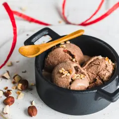 Schokoladiges Nutella-Eis mit 3 Zutaten, garniert mit gehackten Haselnüssen in einer schwarzen Schale, daneben eine Holz-Löffel und rote Bänder.