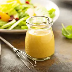 Ein Glas mit gelbem Blattsalat Dressing steht neben einem Schneebesen. Im Hintergrund ein Teller mit frischem Salat.