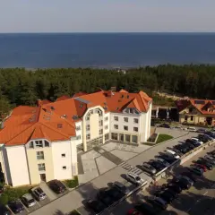 Hotel blisko morza, idealny na wypad z Śląska. Widać parking pełen aut, las i błękitną wodę.