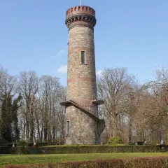 Der Toelleturm in Wuppertal, ein steinerner Aussichtsturm, ragt vor kahlen Bäumen und blauem Himmel auf. Eine Wendeltreppe führt zur Aussichtsplattform.