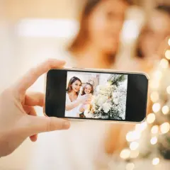 Telefon uchwycił uśmiechnięte mamy i córkę podczas świątecznej sesji w domu. W tle migoczą lampki choinkowe.