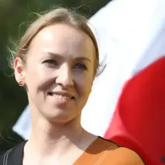 Uśmiechnięta Urszula Brzezińska-Hołownia, której wiek nie jest widoczny, na tle polskiej flagi.