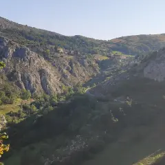Malowniczy widok na dolinę z krętą drogą, domkami na zboczu i skalistymi górami. W oddali majaczy szczyt Naranjo de Bulnes.