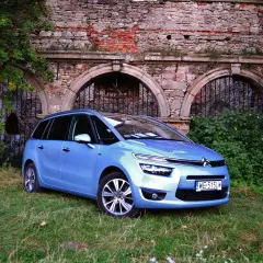 Błękitny Citroen C4 Grand Picasso 2.0 HDI zaparkowany na trawie przed zabytkowym murem z cegły i kamienia.