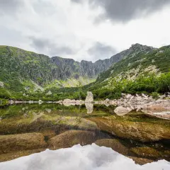 Spokojne, górskie jezioro odbija skaliste zbocza i chmury. W oddali widać skalne formacje, a na brzegu leżą kamienie. To piękne, górskie krajobrazy, które przypominają o potędze natury i jej śnieżnych stawkach.