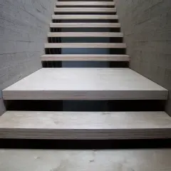 Una scala moderna con gradini in legno compensato, materiale resistente e versatile, fiancheggiata da pareti in cemento grezzo.