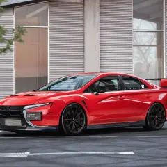 Czerwony Mitsubishi Lancer Evolution XI z czarnymi felgami i spoilerem, zaparkowany przed nowoczesnym budynkiem.