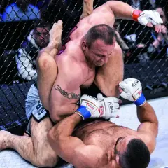 Intensywna walka Pudziana w klatce MMA. Jeden zawodnik klęczy, drugi próbuje go obalić.