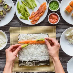 Händer lägger lax och avokado på sushiris för ett sushirecept. Ingredienser som gurka, wasabi och färdig sushi syns också.
