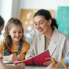 Lehrerin gibt Schülern bei einer Klassenleiterstunde Ideen. Sie schauen gemeinsam in ein Buch, während die Kinder schreiben.