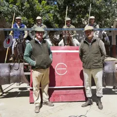 Hombres con gorras y chalecos posan frente a una barrera roja. Uno de ellos, Julio de la Puerta, sonríe. Caballos y equipo de equitación completan la escena.