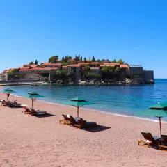 Sonnige Bucht mit rotem Sandstrand und Liegen am Meer in Montenegro. Im Hintergrund die historische Insel Sveti Stefan.