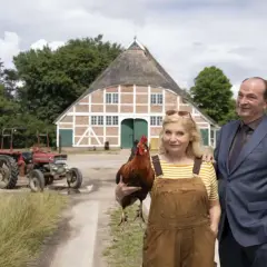Ein Mann im Anzug und eine Frau mit Latzhose und Huhn stehen vor einem Bauernhaus. Im **weiten Land** grasen Schafe, ein Traktor steht bereit.