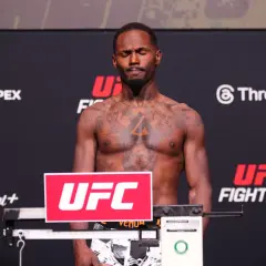 A fighter with tattoos stands on a scale, eyes closed, preparing for weigh-ins. The scale's weight brackets are visible.