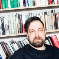 Man med skägg och blå ögon sitter framför bokhyllor. Hans gestaltning, som en person som älskar böcker, framhävs av den rikliga samlingen.