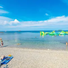 Słoneczna plaża Bijela Uvala z kamyczkami, turkusową wodą i dmuchanymi atrakcjami wodnymi. Ludzie odpoczywają i kąpią się.
