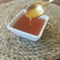 Una gota de salsa agridulce cae de una cuchara dorada a un cuenco blanco sobre una estera tejida.