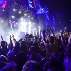 Tłum ludzi z rękami w górze na koncercie, scena z efektami świetlnymi i dymem.