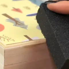 Aplicando pegamento para saber como pegar papel en madera, con una esponja sobre una caja decorada con hojas.