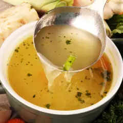 Un cucharón sirve un caldo depurativo, rodeado de verduras frescas como apio, zanahoria y ajo.