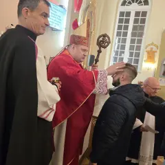 Biskup w czerwonej szacie udziela sakramentu bierzmowania młodemu mężczyźnie w kościele.