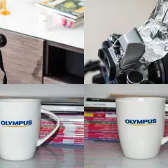 Aparat Olympus z lampą błyskową i dyfuzorem z folii aluminiowej, kubek Olympus na tle stosu magazynów.