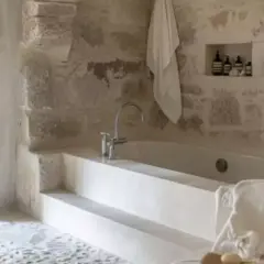 Une **salle de bain méditerranéenne** avec baignoire en pierre brute, robinetterie moderne et serviette blanche suspendue.