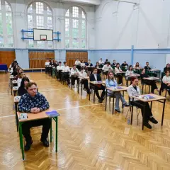 Uczniowie siedzą w ławkach w sali gimnastycznej, zdając egzamin. To typowy widok, gdy młodzi ludzie w wieku ok. 18 lat zdają maturę.