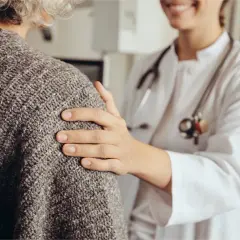 Une doctoresse réconforte une patiente âgée, lui posant une main sur l'épaule. Elle rassure la patiente qui a peur de se faire pipi dessus.