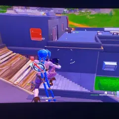 Postać z niebieskimi warkoczami w grze Fortnite buduje drewnianą rampę, celując w budynek. Opis gry Fortnite.