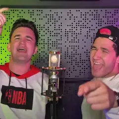 Dwóch mężczyzn śpiewa do mikrofonu. Jeden ma na sobie bluzę z logo NBA, drugi czapkę z daszkiem. W tle widać panel akustyczny.