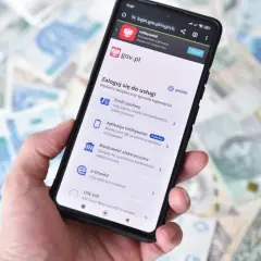 Dłoń trzyma smartfon z ekranem logowania do serwisu gov.pl. Dostępne opcje to m.in. Profil zaufany przez bankowość elektroniczną.