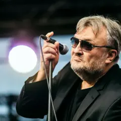 Charyzmatyczny wokalista w okularach przeciwsłonecznych śpiewa do mikrofonu. W tle światła sceniczne, jakby z budki suflera.