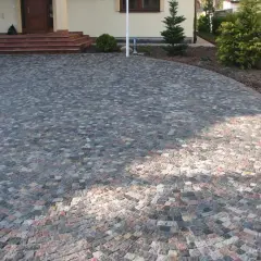 Piękny podjazd z kamienia, ułożony w łuk, prowadzi do drzwi domu. Wokół rosną drzewka i krzewy.