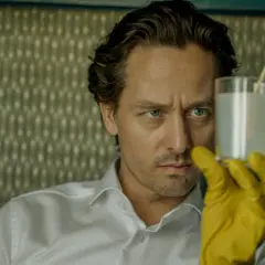 Ein Mann mit blauen Augen und gelben Handschuhen hält ein Glas mit weißer Flüssigkeit. Er ist Teil der "Achtsam Morden Reihe".