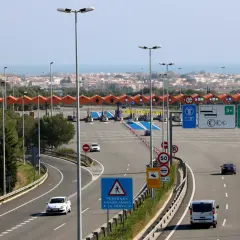 Carriles de peaje con barreras y señales de tráfico. Un coche blanco se acerca al peaje garraf, con la ciudad y el mar al fondo.