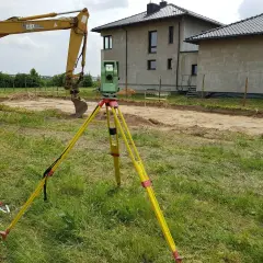 Geodeta z niwelatorem na statywie na placu budowy obok koparki i domów.